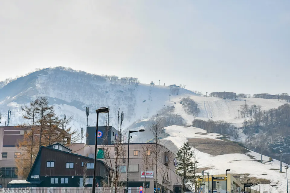 A scenic view of a snowy mountain with ski tracks, set behind a row of buildings. The sky is clear, and a few leafless trees are scattered in the foreground.