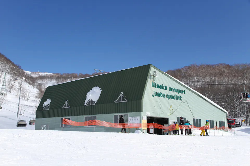 Skiers gather outside a green building labeled "Niseko Annupuri Jumbo Quad #1" at a snowy ski resort. The sky is clear and blue, surrounded by snow-covered hills. A ski lift is partially visible on the right.