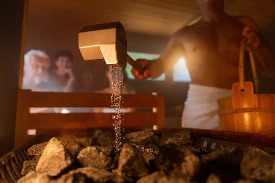 A person pours water from a ladle onto hot stones in a sauna, creating steam. Others are seated in the background, blurred. The setting is warm and wooden, with a cozy atmosphere.