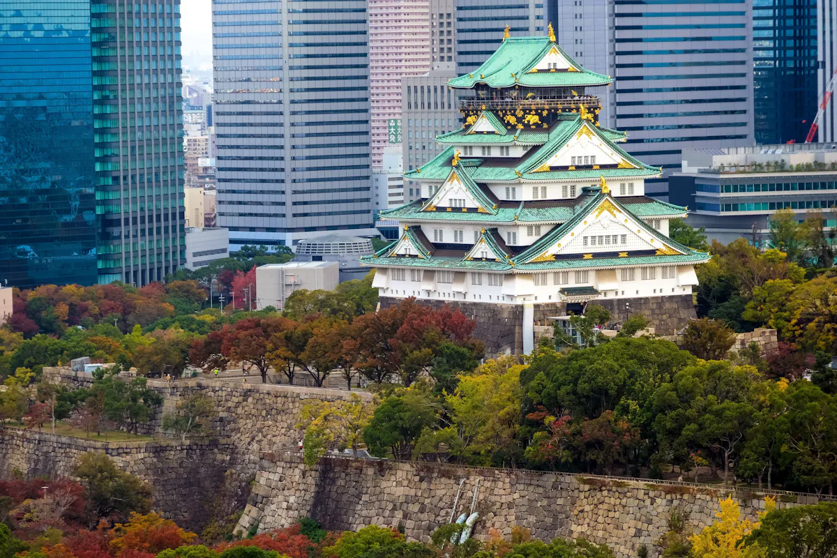 Osaka Castle surrounded by colorful autumn trees, set against a backdrop of modern skyscrapers. The castle's traditional architecture features green and gold-tipped roofs, creating a striking contrast with the urban skyline.