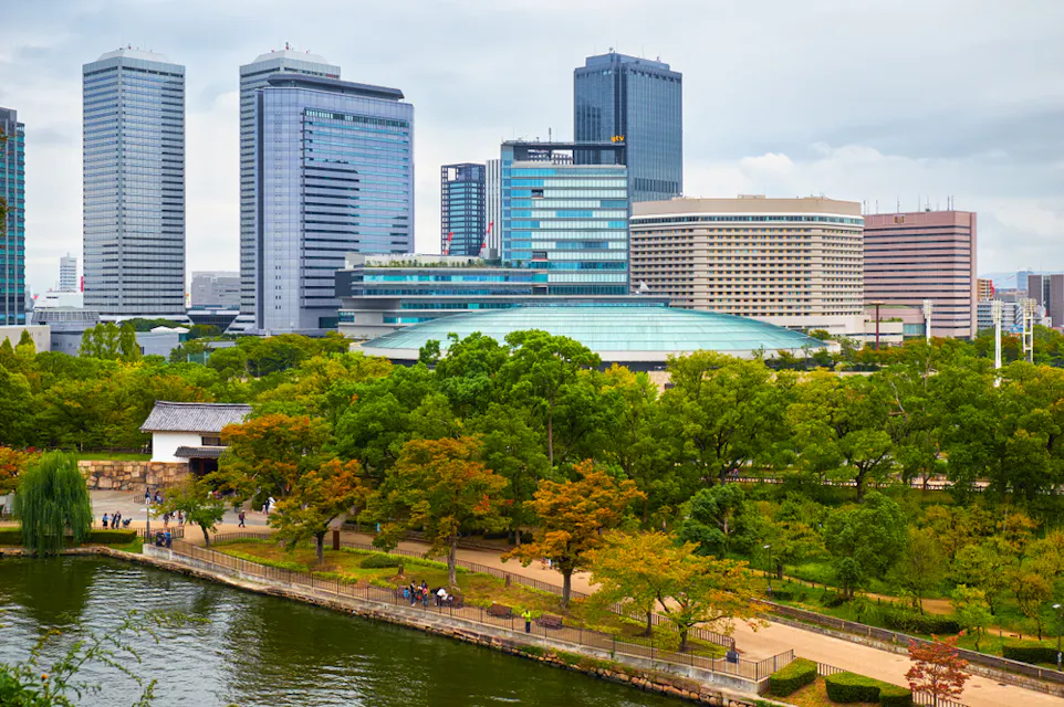 A cityscape view featuring a lush park with trees and a pathway alongside a river in the foreground. Tall modern buildings and skyscrapers are visible in the background under a cloudy sky.