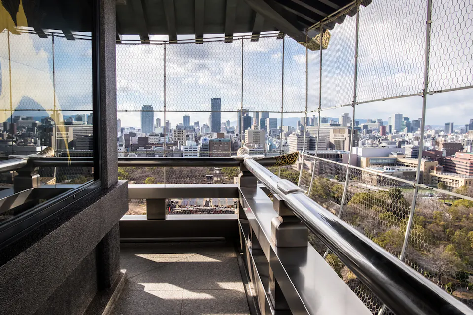 A high vantage point view from an observation deck overlooking a cityscape with tall buildings and a vast sky. The deck is enclosed with wire mesh fencing and features reflective glass panels, revealing trees and urban infrastructure below.