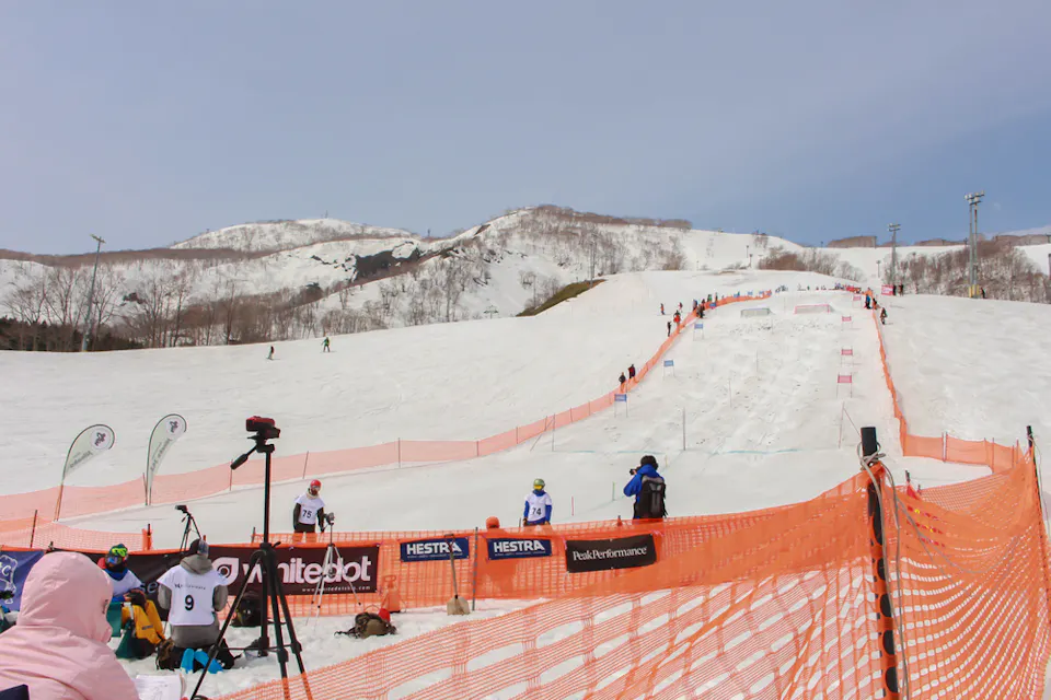 A snowy ski slope with orange safety fencing and a mogul course. Skiers in numbered bibs are preparing at the bottom, with a few spectators and trainers nearby. Snow-covered hills and a clear sky are in the background.