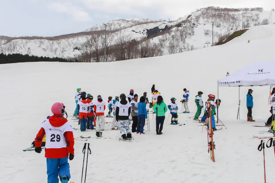 A group of skiers wearing numbered bibs gather at the bottom of a snowy slope under a cloudy sky. They appear to be preparing for a ski event, with additional people in colorful jackets standing nearby. Skis are propped up in the snow.