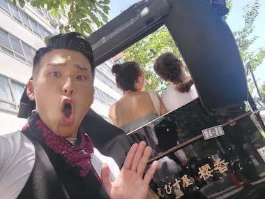 A man with a surprised expression poses in front of a traditional rickshaw carrying two women with braided hair, seen from behind, on a sunny day beside a modern building and leafy trees.