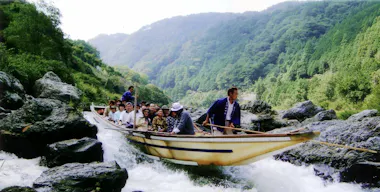 A group of people ride in a wooden boat navigated by a guide through whitewater rapids, surrounded by lush green hills and rocky terrain under a bright sky.