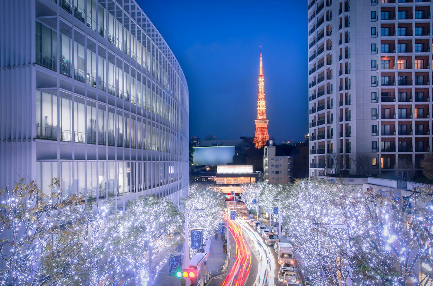 A cityscape at night features a brightly lit street with buildings adorned in white lights. In the background, an illuminated tower glows against the dark sky. Traffic creates light trails, adding to the vibrant urban scene.