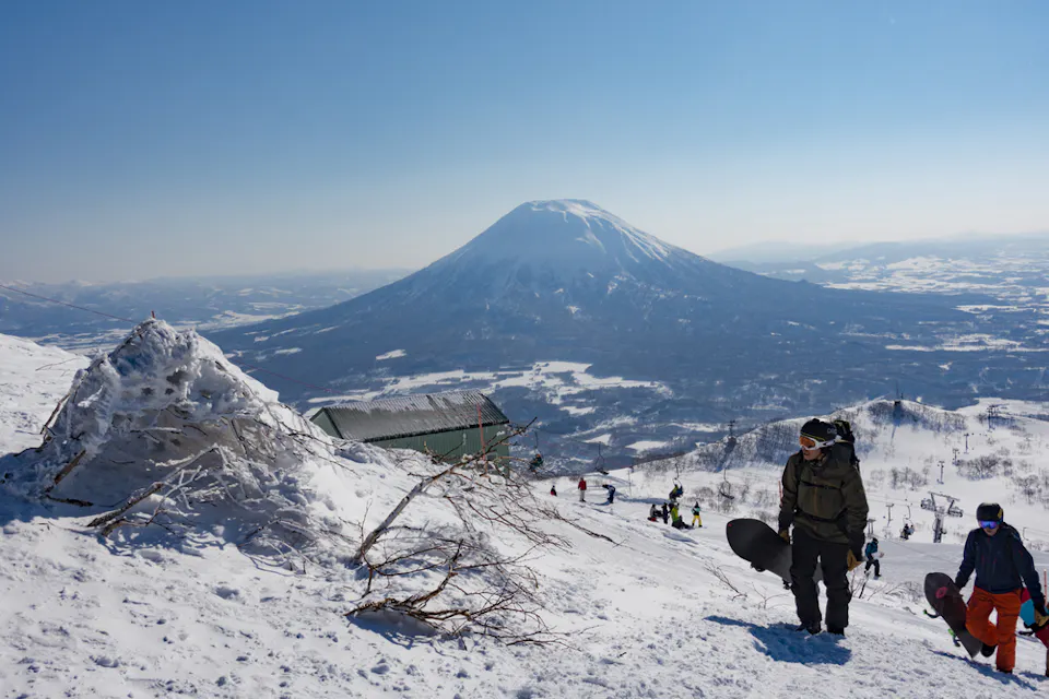 Two people in winter gear walk up a snowy mountain slope, carrying snowboards. In the background, a large, majestic mountain stands under a clear blue sky. Other snow-covered hills and trees are visible in the landscape.