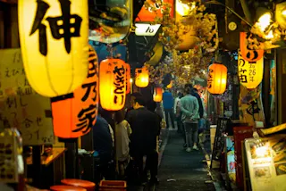 A narrow, bustling alleyway in Japan is lit by colorful paper lanterns with Japanese writing. People walk beneath the lanterns, and restaurant signs and cherry blossom decorations line the vibrant street.