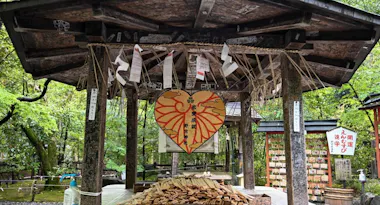 A traditional Japanese wooden pavilion with a large, heart-shaped ema plaque decorated with red patterns and Japanese writing, surrounded by smaller wooden plaques and paper fortunes. Lush greenery is visible in the background.