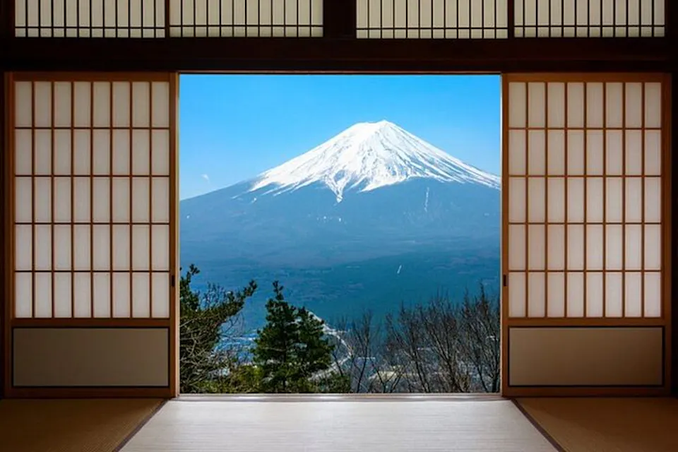 A view of Mount Fuji framed by traditional Japanese shoji screens. The scene captures the snow-capped peak of the mountain under a clear blue sky, seen from the interior of a room with wooden elements.