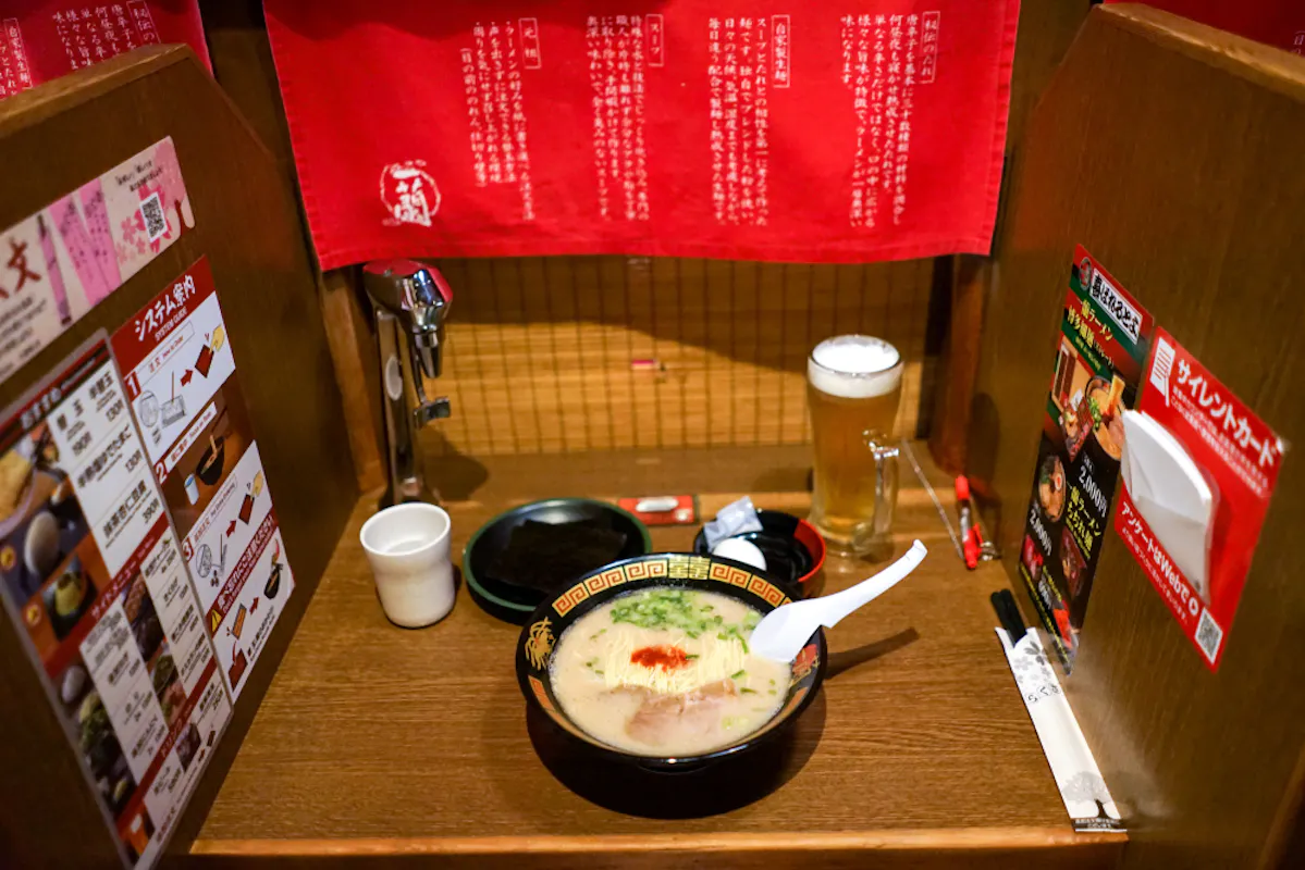 A bowl of ramen with a white spoon on a counter, accompanied by a glass of beer and a cup of water. The booth is decorated with red curtains and Japanese menus on the side walls.