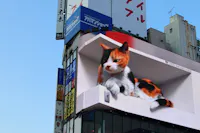 A large 3D digital billboard displays a realistic calico cat peeking out from a box, above a busy building with various colorful signs and advertisements in a city.