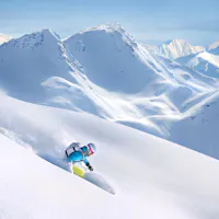 Heli-Skiing A person skiing down a pristine, powdery slope in a mountainous landscape. They are wearing a blue jacket and a helmet. Snow-covered peaks are visible in the background under a clear blue sky.