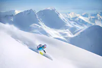 A person skiing down a pristine, powdery slope in a mountainous landscape. They are wearing a blue jacket and a helmet. Snow-covered peaks are visible in the background under a clear blue sky.