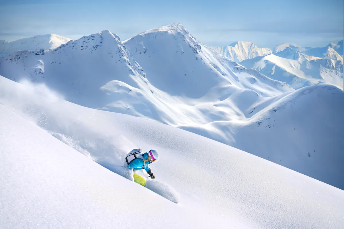 A person skiing down a pristine, powdery slope in a mountainous landscape. They are wearing a blue jacket and a helmet. Snow-covered peaks are visible in the background under a clear blue sky.