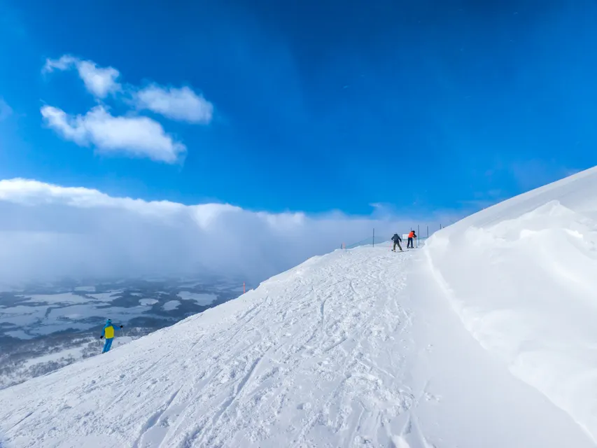 Three skiers descend a snow-covered mountain slope under a vibrant blue sky. The landscape below is partially shrouded in mist, creating a scenic winter vista.