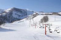 A snowy mountain landscape with a ski lift carrying people up the slope. Skiers are scattered across the snowy terrain, and the sky is clear and blue. The hillside is dotted with leafless trees.