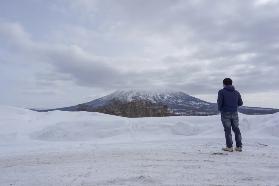 A person in a blue jacket stands on a snowy road, facing a distant snow-covered mountain under a cloudy sky.