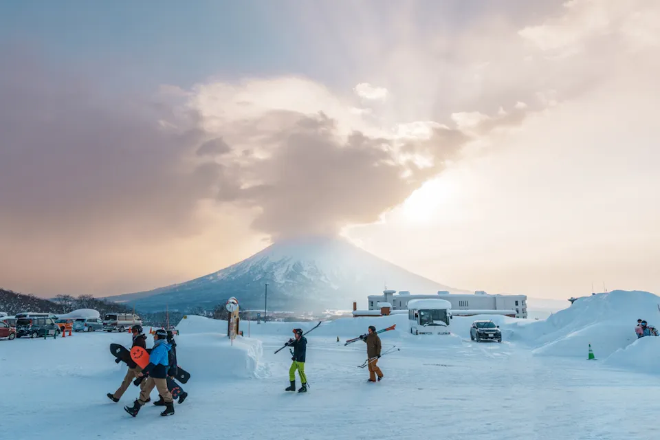 People carrying snowboards and skis walk across a snow-covered landscape with a majestic sunlit mountain in the background. The sky is partly cloudy, and vehicles are parked nearby.