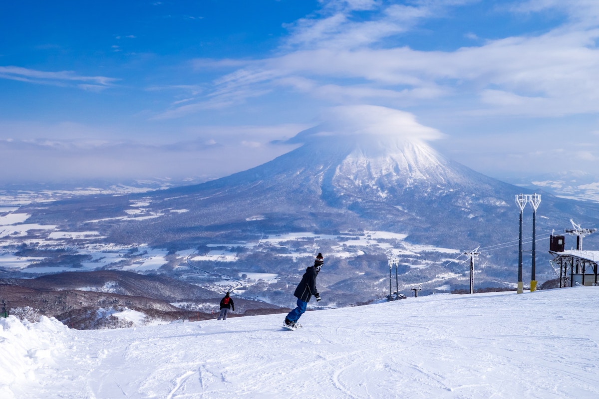 Ski Resort Two snowboarders glide down a snowy slope with a large, cloud-capped mountain in the background under a blue sky. Ski lifts are visible on the right.