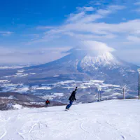 Ski Resort Two snowboarders glide down a snowy slope with a large, cloud-capped mountain in the background under a blue sky. Ski lifts are visible on the right.