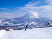 Two snowboarders glide down a snowy slope with a large, cloud-capped mountain in the background under a blue sky. Ski lifts are visible on the right.