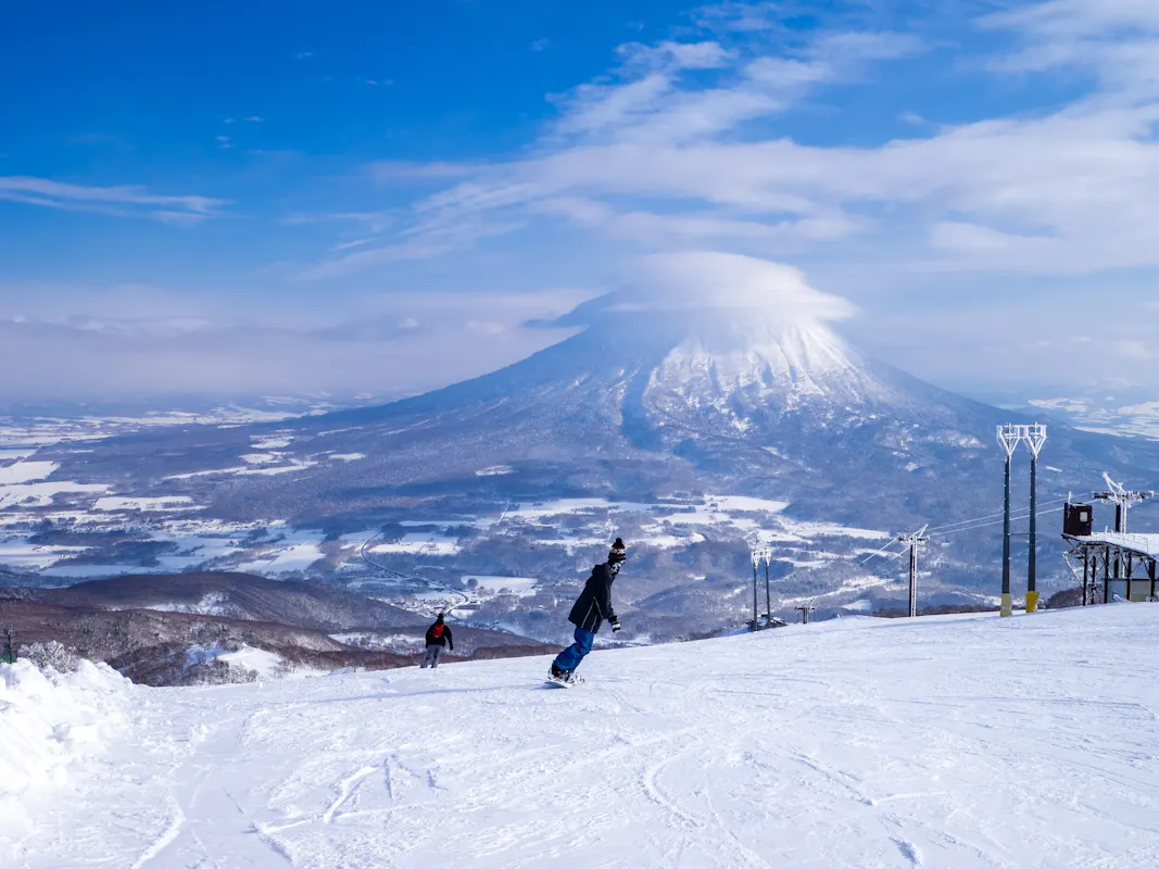Two snowboarders glide down a snowy slope with a large, cloud-capped mountain in the background under a blue sky. Ski lifts are visible on the right.