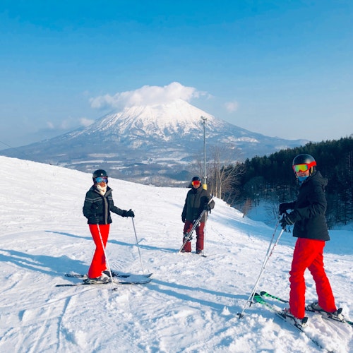 Niseko Hirafu Ski Resort Three people in ski gear, wearing red pants and helmets, stand on a snowy slope with ski poles in hand. A snow-covered mountain and a clear blue sky provide a scenic backdrop. Ski lifts and a line of trees are visible in the background.