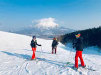 Three people in ski gear, wearing red pants and helmets, stand on a snowy slope with ski poles in hand. A snow-covered mountain and a clear blue sky provide a scenic backdrop. Ski lifts and a line of trees are visible in the background.