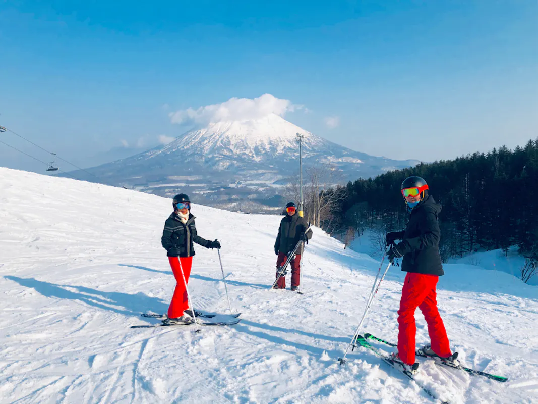 Three people in ski gear, wearing red pants and helmets, stand on a snowy slope with ski poles in hand. A snow-covered mountain and a clear blue sky provide a scenic backdrop. Ski lifts and a line of trees are visible in the background.