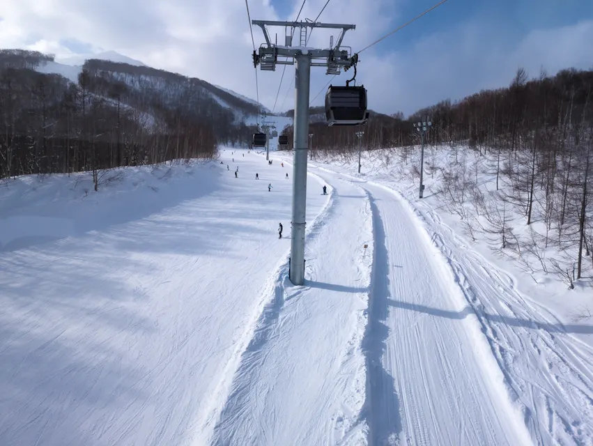 Grand Hirafu Gondola A snowy landscape with a ski lift running parallel to a ski trail. Skiers are visible on the slope, surrounded by snow-covered trees under a clear sky with scattered clouds.