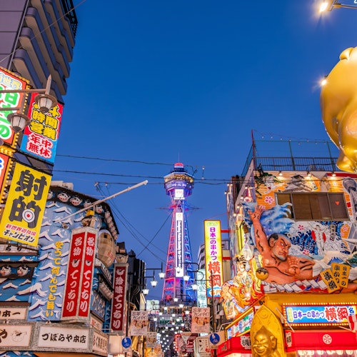 Osaka A vibrant street scene in Osaka, Japan, with colorful neon signs and billboards. The famous Tsutenkaku Tower is visible in the background, illuminated against a clear evening sky. The area is bustling with energy and lively advertising displays.