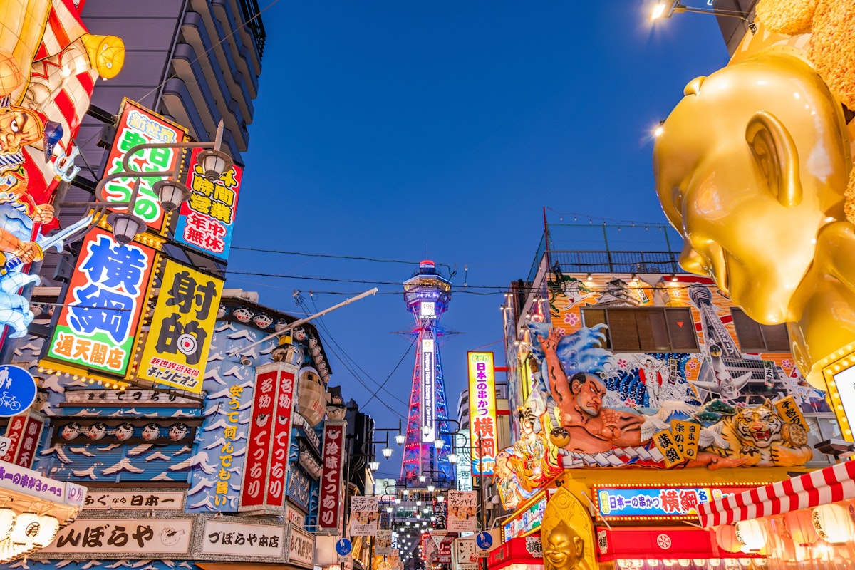 Osaka A vibrant street scene in Osaka, Japan, with colorful neon signs and billboards. The famous Tsutenkaku Tower is visible in the background, illuminated against a clear evening sky. The area is bustling with energy and lively advertising displays.