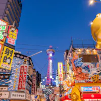 Osaka A vibrant street scene in Osaka, Japan, with colorful neon signs and billboards. The famous Tsutenkaku Tower is visible in the background, illuminated against a clear evening sky. The area is bustling with energy and lively advertising displays.