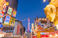 A vibrant street scene in Osaka, Japan, with colorful neon signs and billboards. The famous Tsutenkaku Tower is visible in the background, illuminated against a clear evening sky. The area is bustling with energy and lively advertising displays.