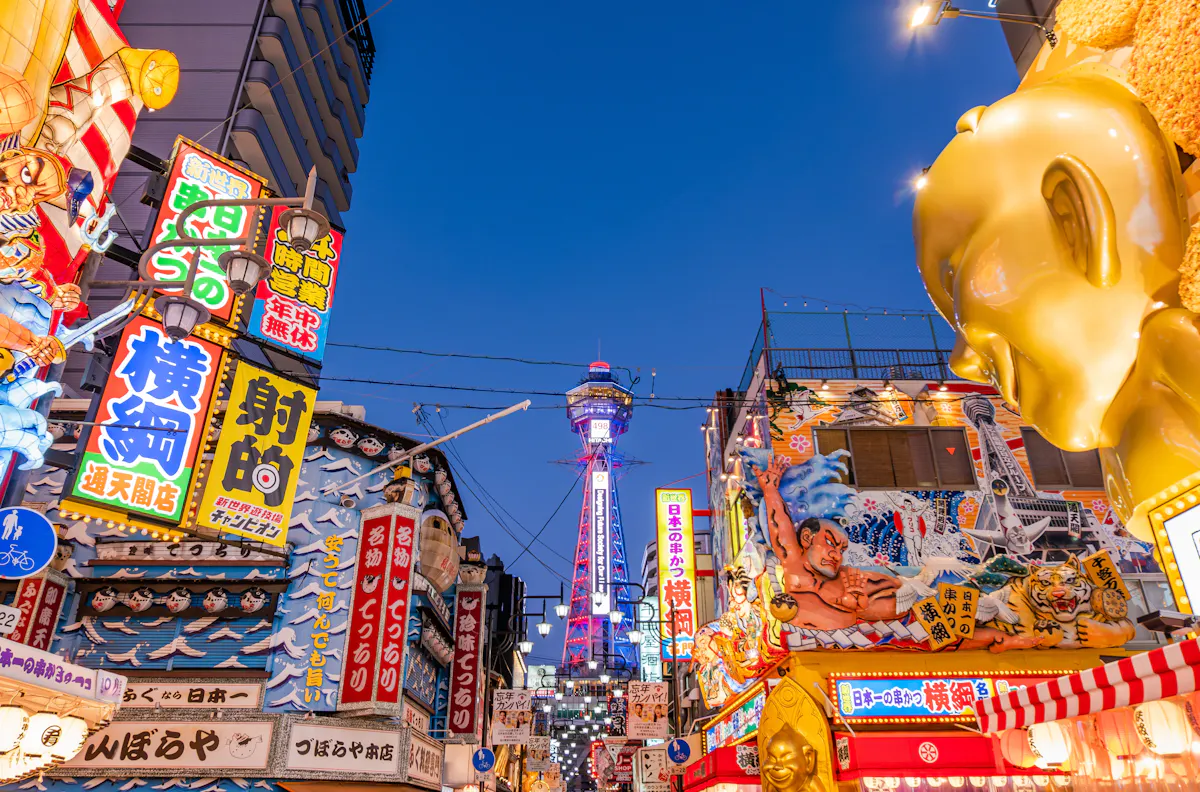 A vibrant street scene in Osaka, Japan, with colorful neon signs and billboards. The famous Tsutenkaku Tower is visible in the background, illuminated against a clear evening sky. The area is bustling with energy and lively advertising displays.