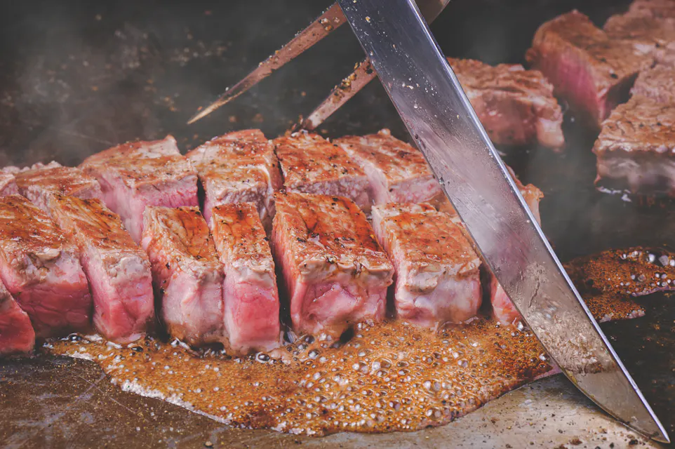 A close-up of a sizzling steak being sliced on a grill. The meat is juicy with a seared crust, showing varying shades of pink and brown. Steam rises as it cooks, and juices pool around the slices, highlighting the steak's tenderness.