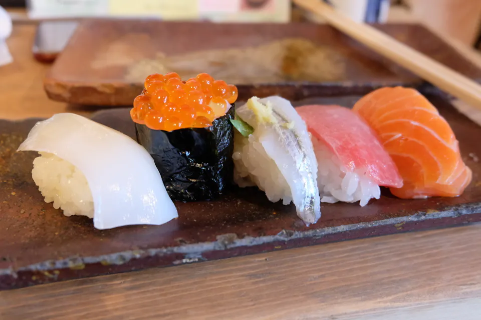 A platter of assorted sushi on a rectangular plate, featuring squid, salmon roe, mackerel, tuna, and salmon pieces, all neatly arranged on vinegared rice. Chopsticks are visible in the background.
