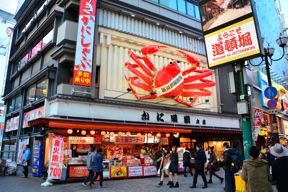 Bustling street scene in Dotonbori, Osaka, with a large crab sign on a building. People walk past bright signs and colorful storefronts in the lively shopping area.