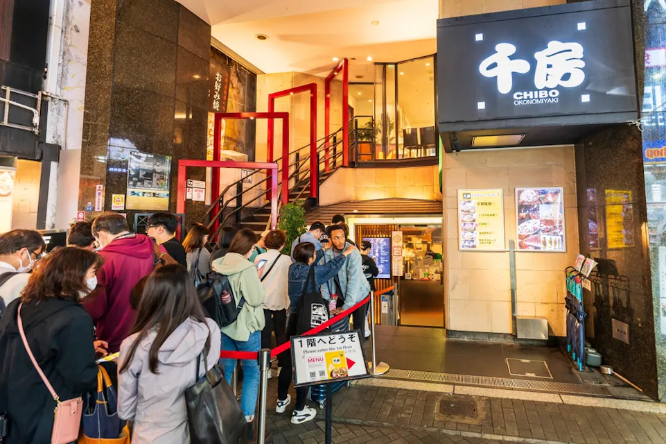 A line of people wait outside a restaurant with a lit sign, "CHIBO OKONOMIYAKI," and bright interior lighting. Some individuals wear masks. The entrance features red railings and a menu display. The surrounding area appears lively and urban.