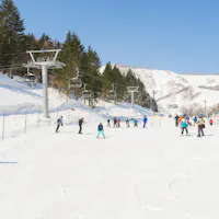 Grand Hirafu People skiing and snowboarding on a snowy slope at a ski resort. The area is surrounded by trees and mountains under a clear blue sky. A chairlift is visible on the left, and skiers are dispersed across the slope near a building on the right.