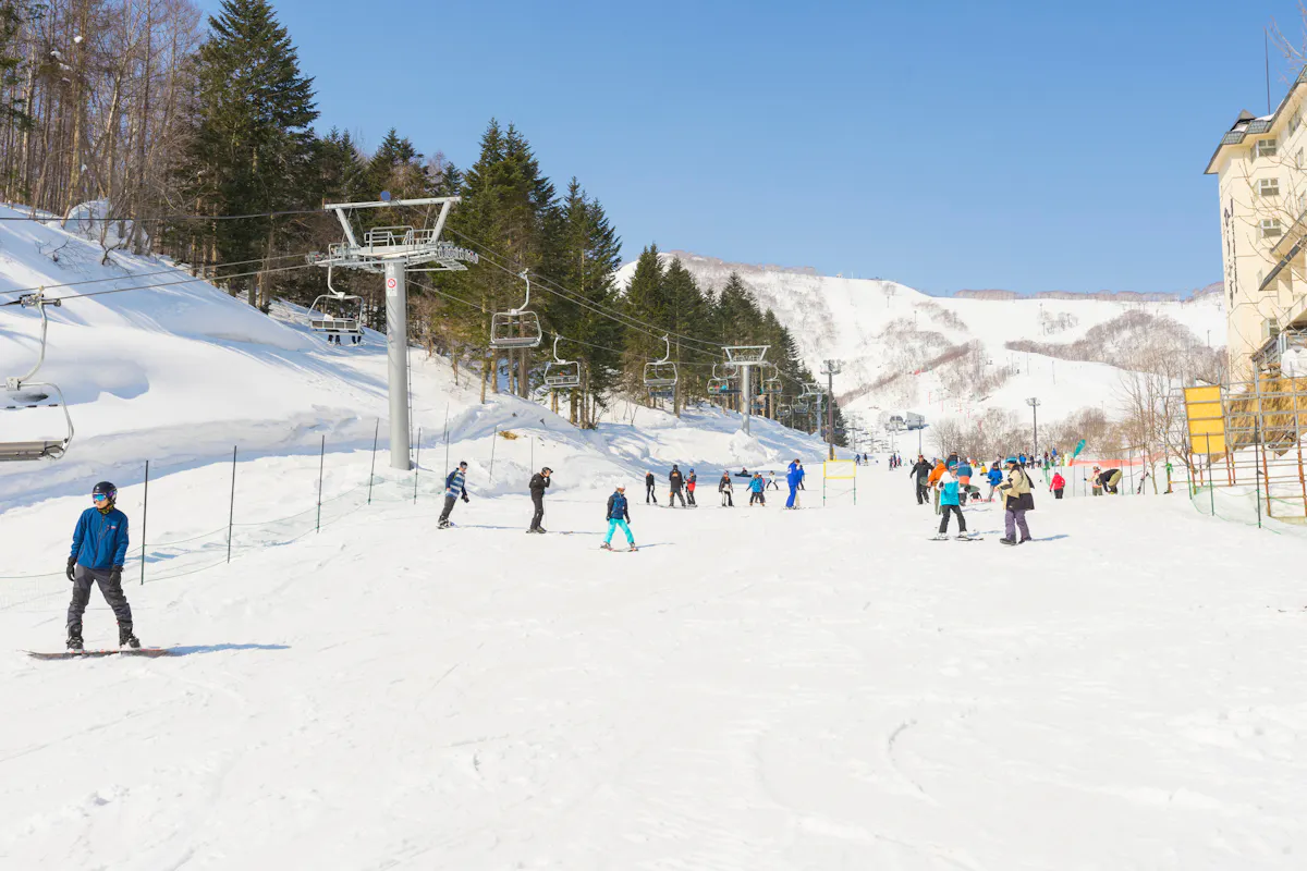 Grand Hirafu People skiing and snowboarding on a snowy slope at a ski resort. The area is surrounded by trees and mountains under a clear blue sky. A chairlift is visible on the left, and skiers are dispersed across the slope near a building on the right.