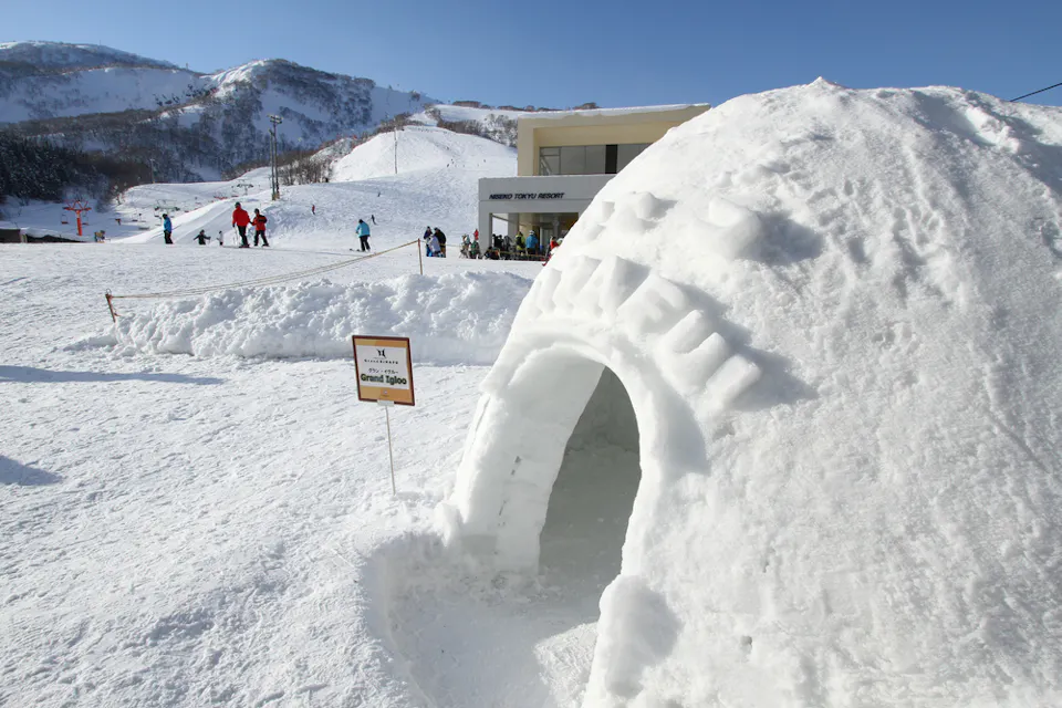 A large snow igloo on a snowy landscape with mountains in the background. People are gathered near a sign beside the igloo, and a modern building is visible nearby. The sky is clear and blue.