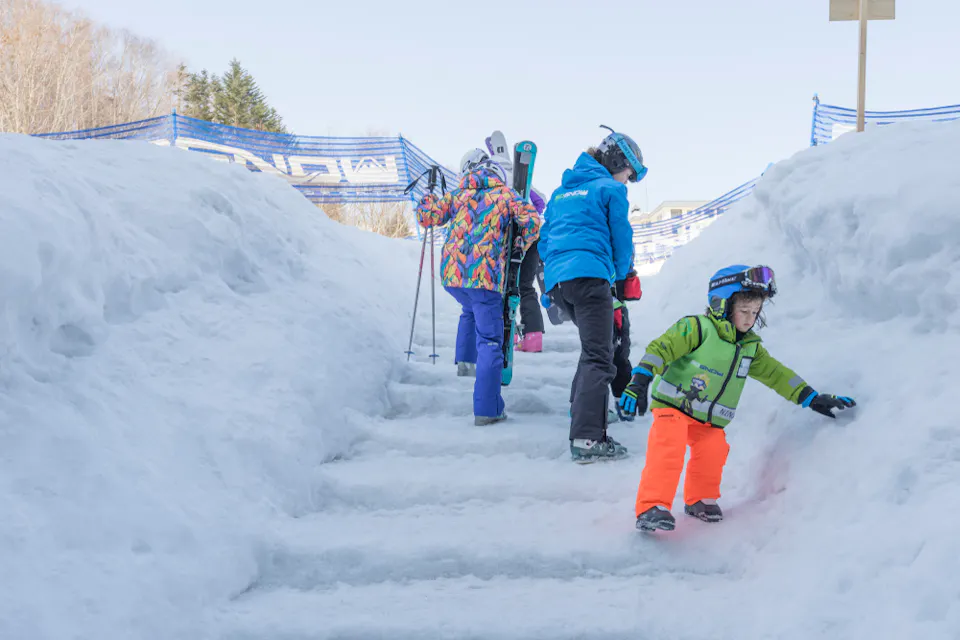 Three children are walking up snowy steps while wearing colorful winter clothing and ski gear. Two carry skis, and one is dressed in a green jacket and orange pants. Snow piles and a blue fence line the path, with trees in the background.