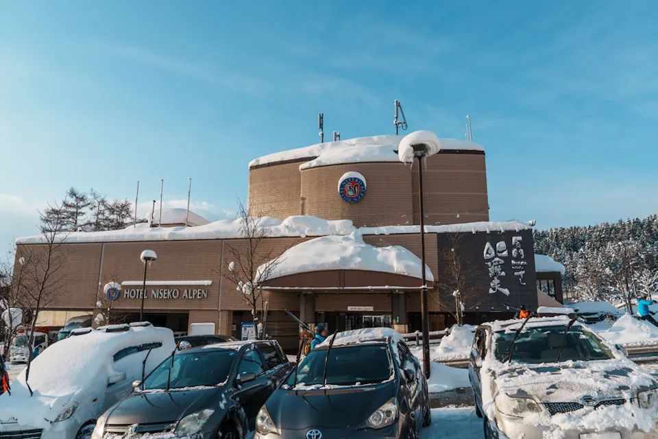 A hotel building in a snowy landscape with cars parked in front, covered in snow. The sky is clear and blue, and the hotel sign reads "Hotel Niseko Alpen." Snow blankets the ground and trees.