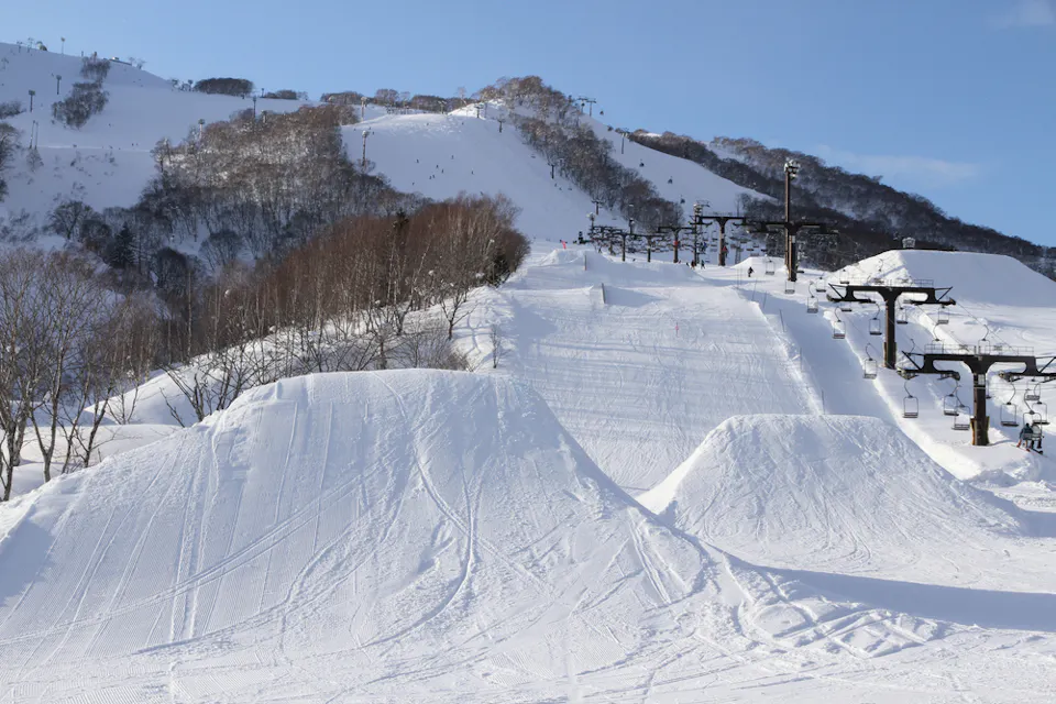 A snowy ski resort with clear blue skies. The foreground features snow-covered slopes with ski jumps and tracks. Ski lifts with chairs are on the right side, and tree-covered hills rise in the background.