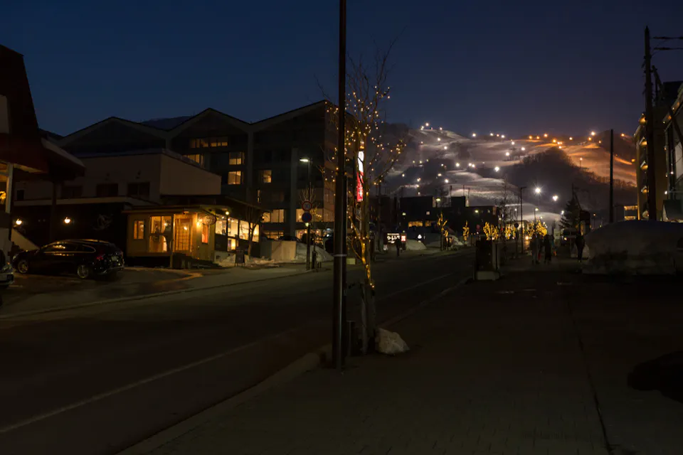 A nighttime street scene in a ski town, with warmly lit buildings on the left. A distant ski slope is brightly illuminated, with lights creating a trail pattern on the snow-covered hill. Snow is visible on the roadside.