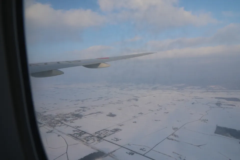View from an airplane window showing a snowy landscape below with patches of fields and buildings. The wing of the airplane is visible against a backdrop of a partly cloudy sky.