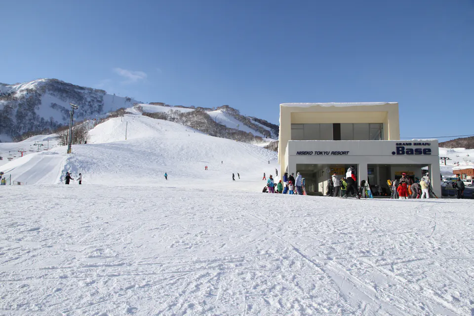 Ski resort scene with a snowy landscape and people skiing and snowboarding. A modern building labeled "Niseko Tokyu Resort .Base" stands on the right. Mountains and ski lifts are in the background under a clear blue sky.